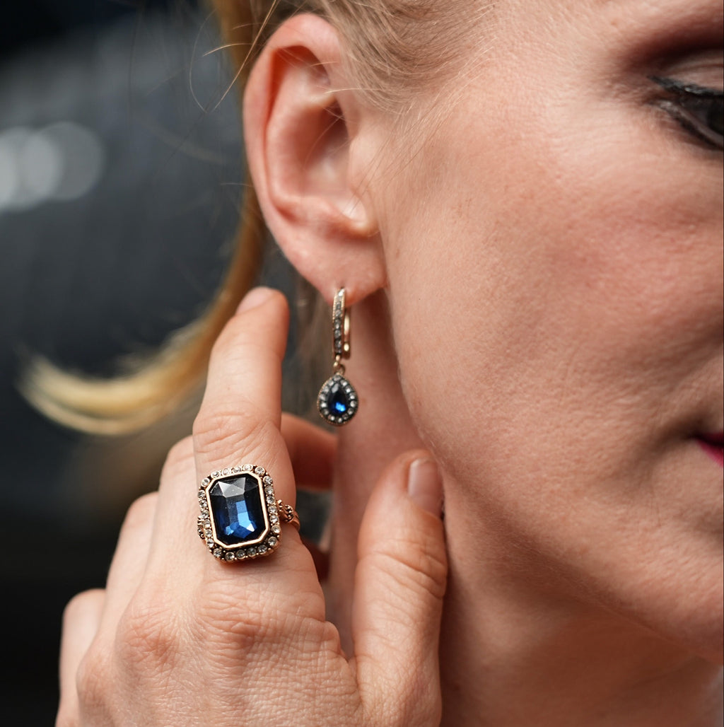 Close-up of a woman wearing a blue gemstone ring and earring, with a blurred background.