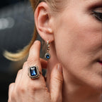 Close-up of a woman wearing a blue gemstone ring and earring, with a blurred background.