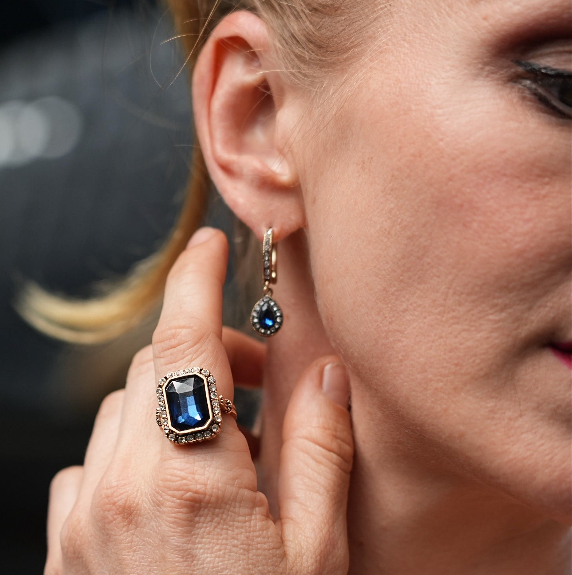 Close-up of a woman wearing a blue gemstone ring and earring, with a blurred background.