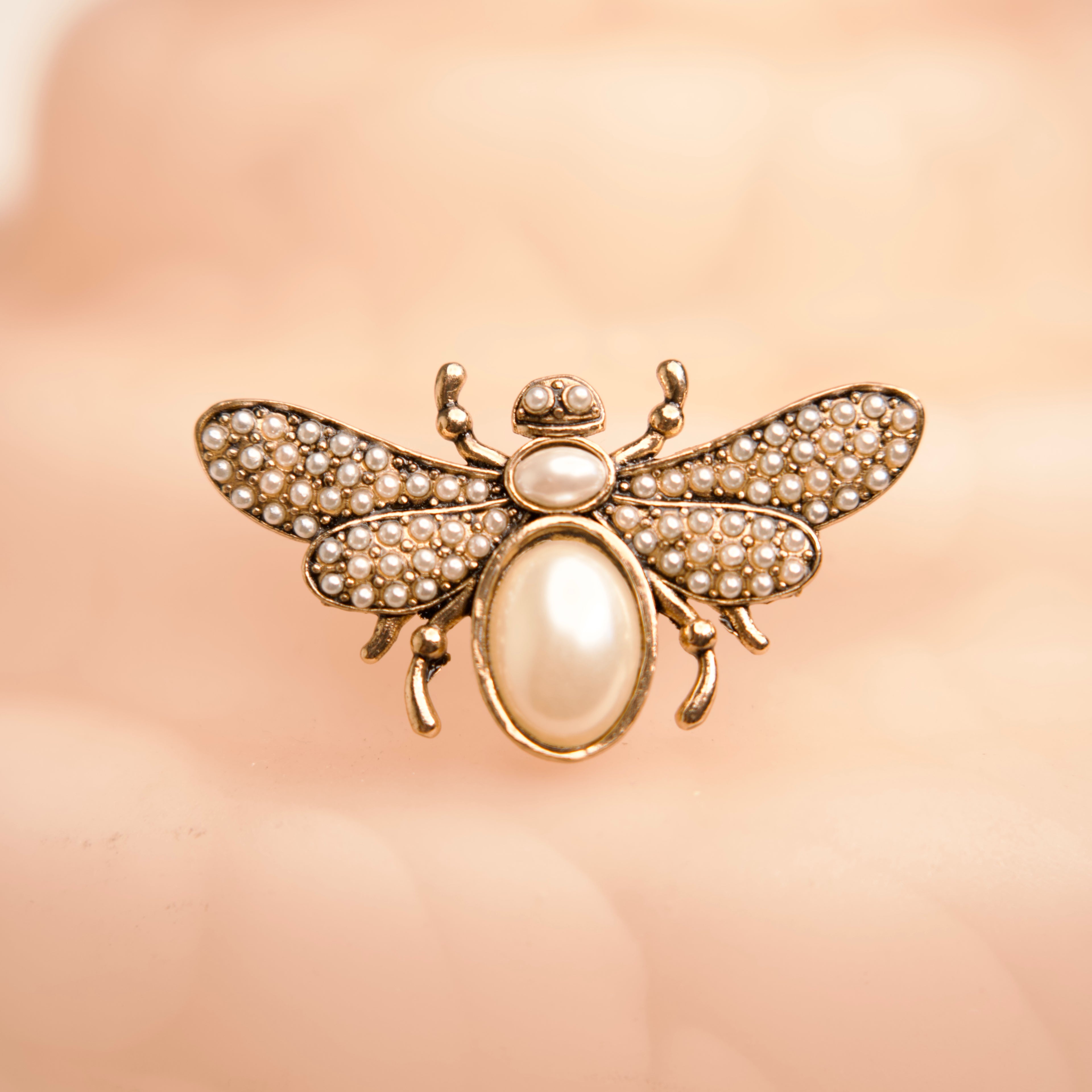 Gold bee-shaped brooch with pearl accents on a soft pink background