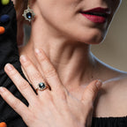Woman wearing a black dress with colorful pom-poms, holding her hand near her face, with a close-up of her jewelry.