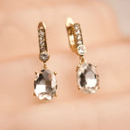 Gold earrings with clear gemstones on a soft pink background