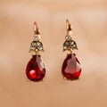 Pair of red teardrop earrings with gold and gemstone accents on a beige background