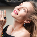 Woman wearing a black top with colorful pom-poms, holding a ring with a blue gemstone.
