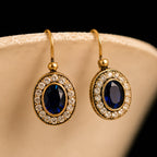 A pair of gold drop earrings with oval-cut dark blue center stones surrounded by a halo of small clear stones, displayed on a beige jewelry stand against a black background.